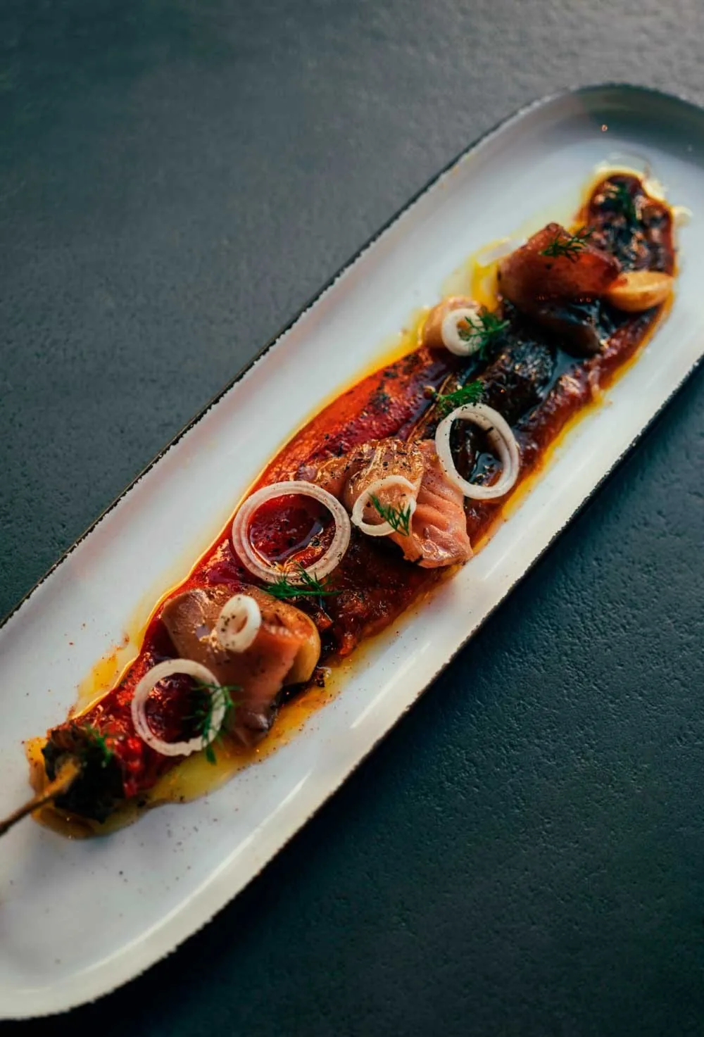 A long white plate of sliced fish topped with tomato and onion on a dark teal surface