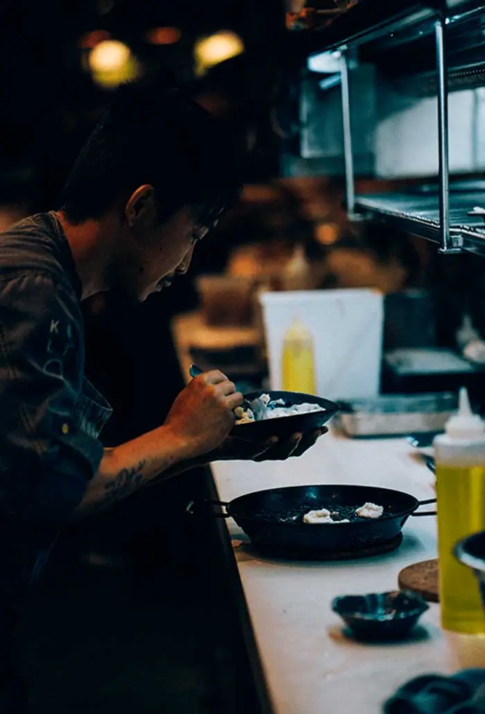 Chef plating food from a small pan at the kitchen counter, dim lighting