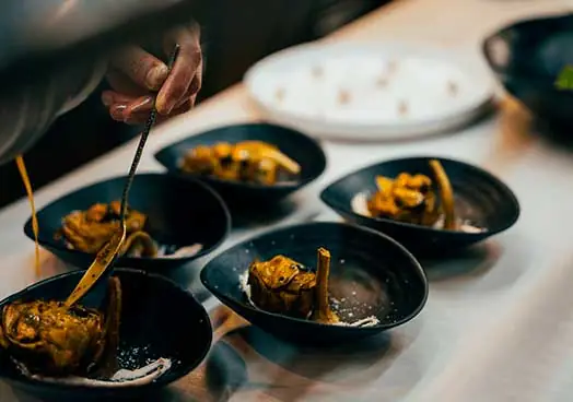 Hand plating small dishes of a charred vegetable in dark bowls on a kitchen counter