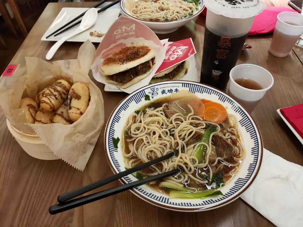 A bowl of noodle soup beside steamed buns and drinks on a table