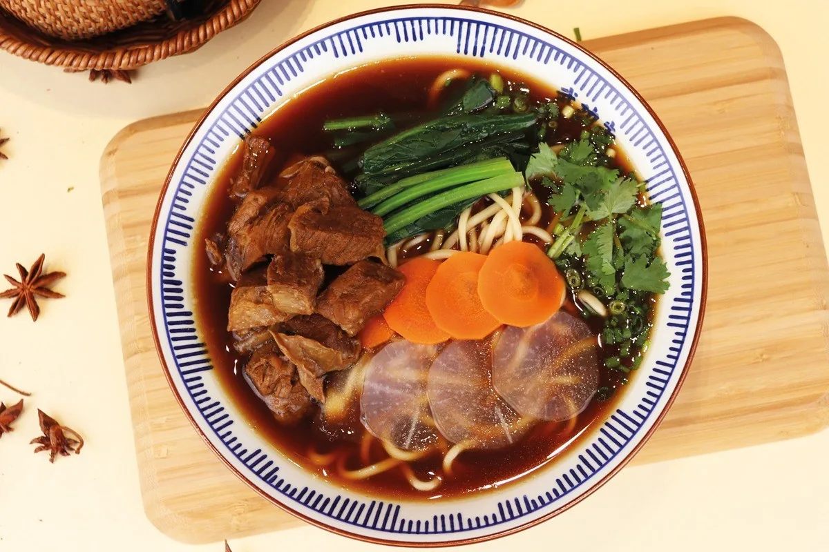 A blue and white bowl of noodles in dark broth with chunks of braised beef, greens, carrot and radish slices, seen from above