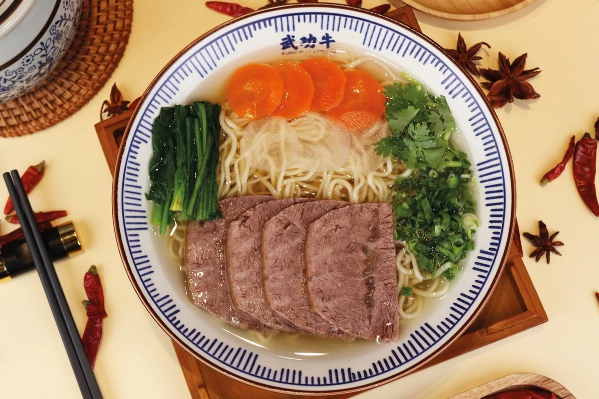 A blue and white bowl of noodles in clear broth with sliced beef, greens and carrot on a wooden board