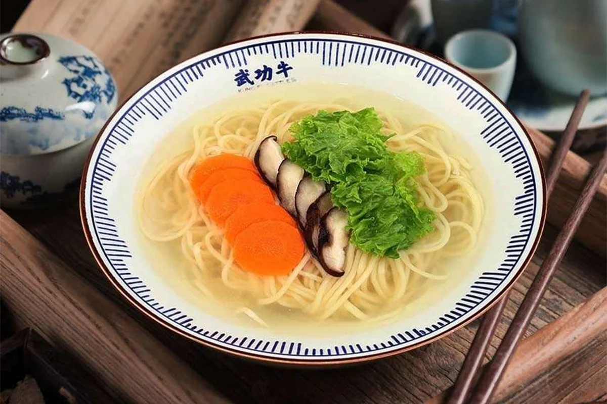 A blue and white bowl of noodles in clear broth with carrot slices, mushroom slices and lettuce