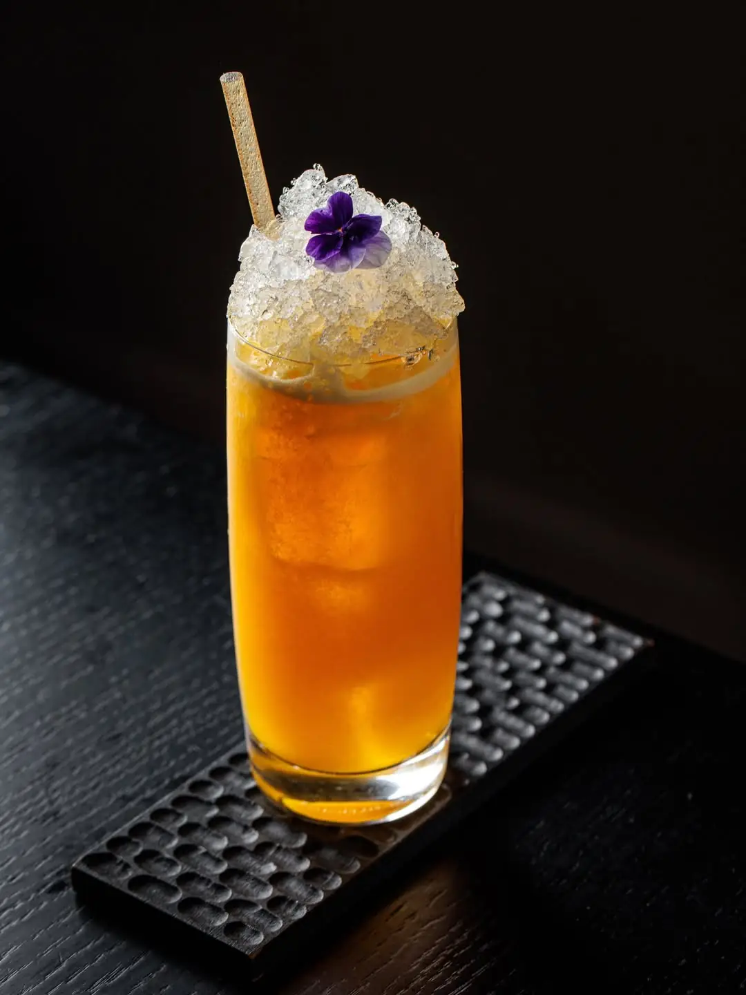 Tall orange cocktail with crushed ice and a purple flower garnish