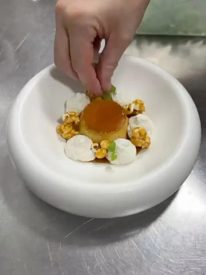 Hand garnishing a caramel flan surrounded by cream and popcorn