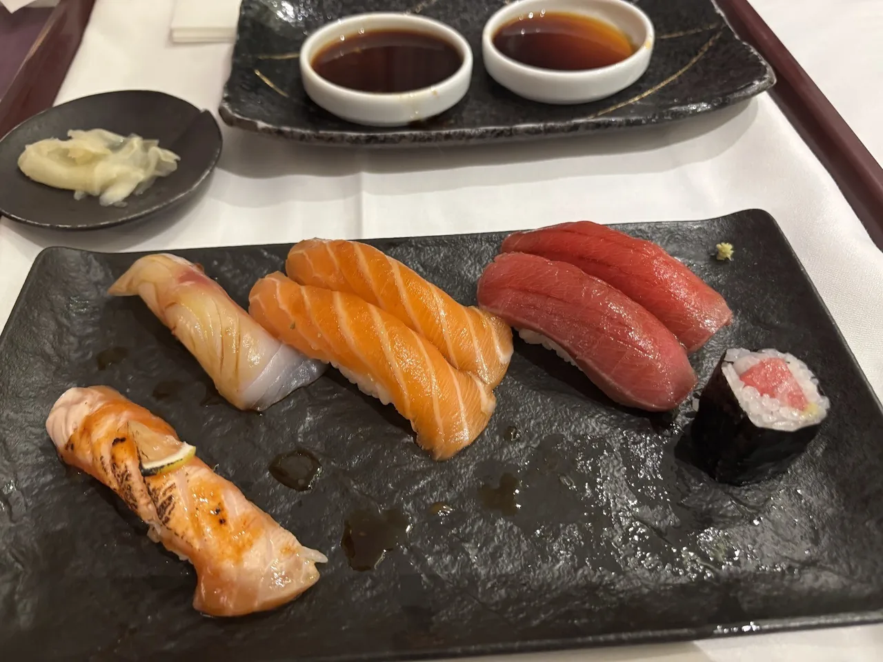 An assortment of nigiri on a black slate plate with small dishes of soy sauce and pickled ginger