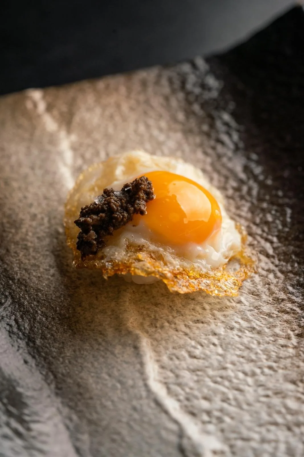 A small bite topped with a quail egg yolk and dark garnish on a textured plate