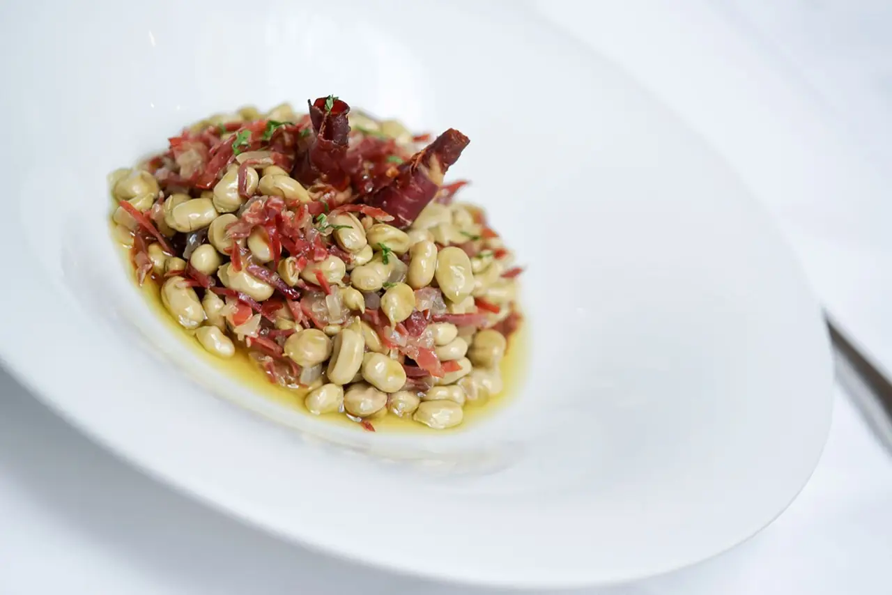 Sauteed broad beans with shredded cured ham and crisp ham crackling in olive oil, plated in a white bowl