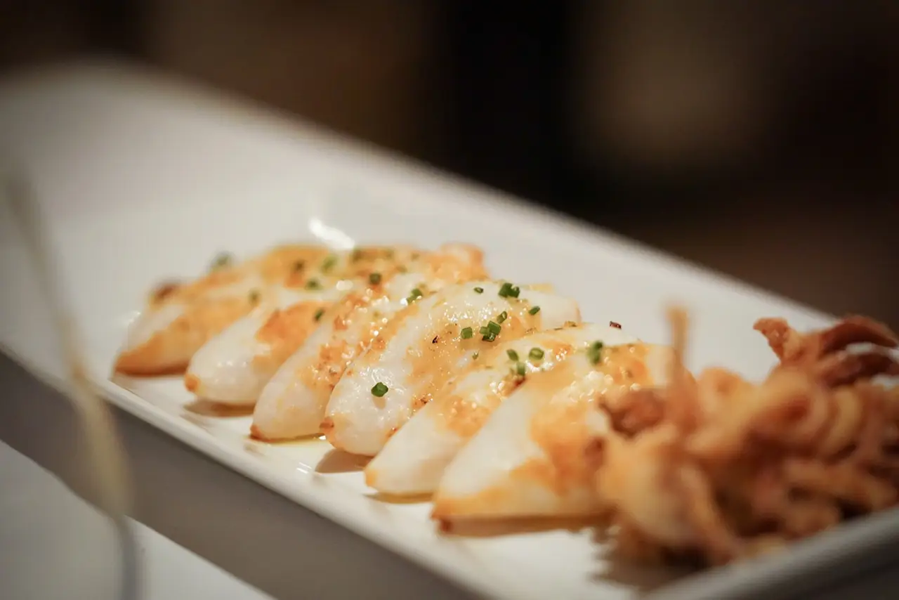 Row of grilled squid pieces glazed and sprinkled with chives on a rectangular white platter, with fried tentacles at the end