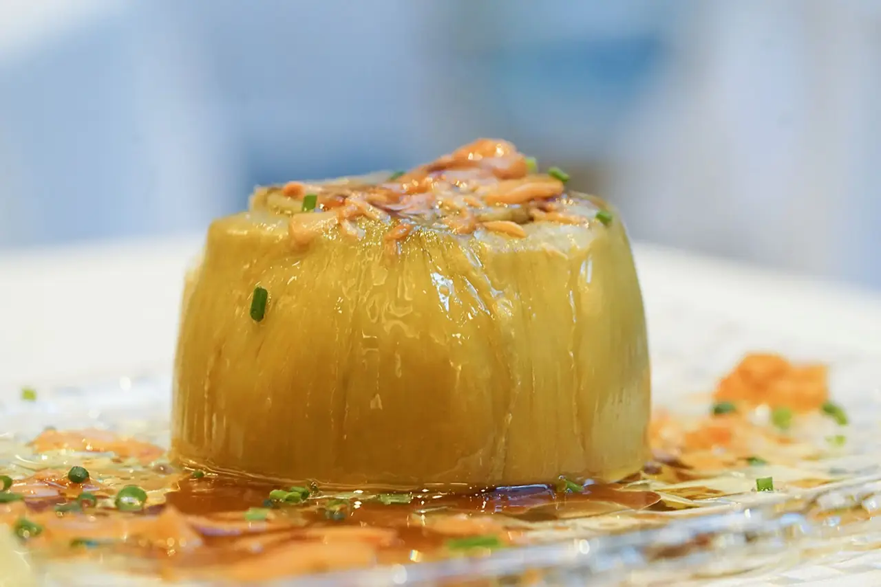 Whole braised artichoke heart topped with shredded jamon and chives, sitting in a pool of broth on a glass plate