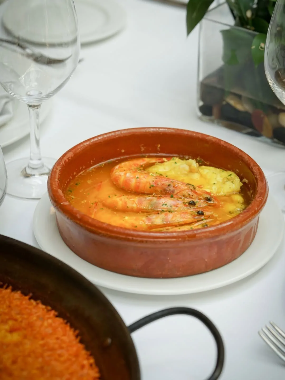 Prawns in an orange sauce in a clay dish beside a paella pan