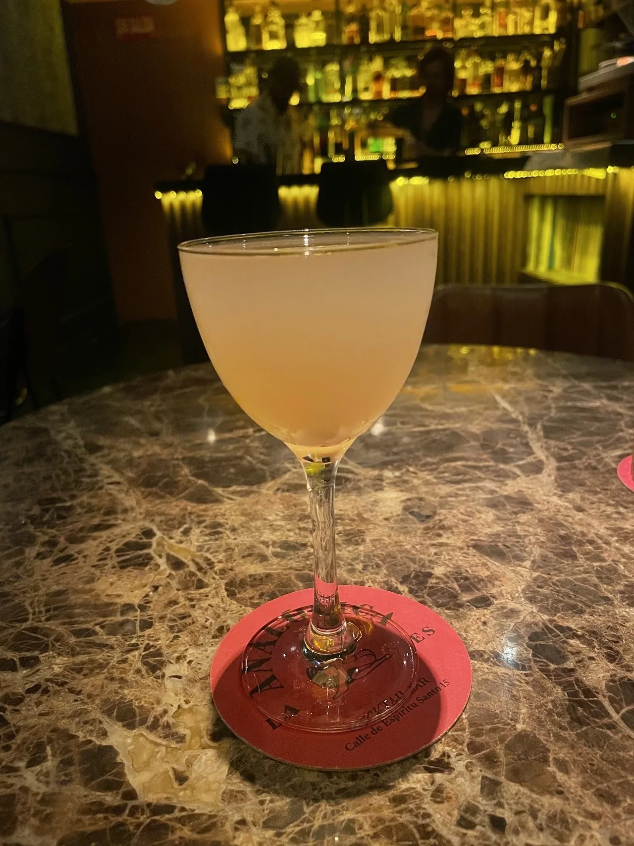 A pale cocktail in a coupe glass on a coaster on a marble bar top