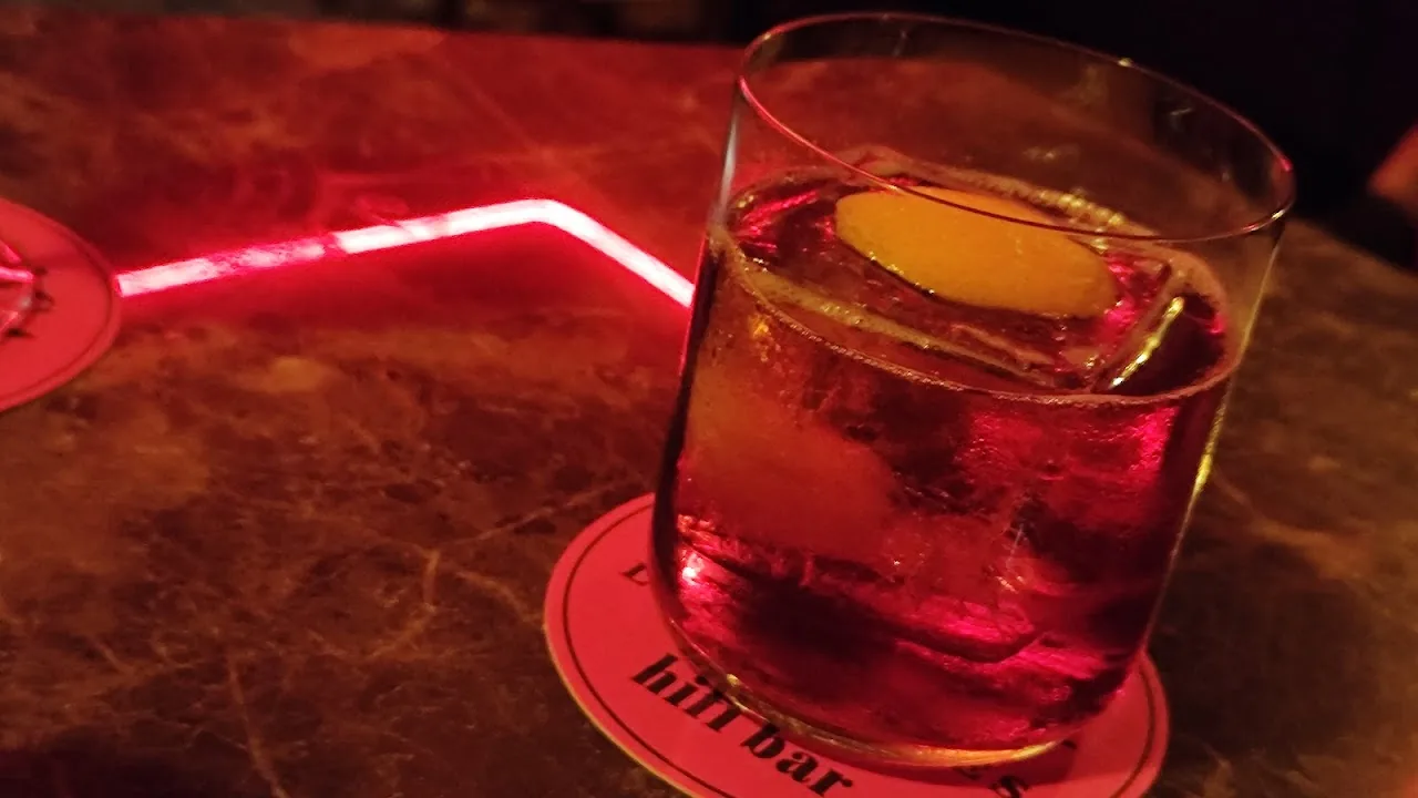 A rocks glass cocktail with ice on a bar lit by red neon light