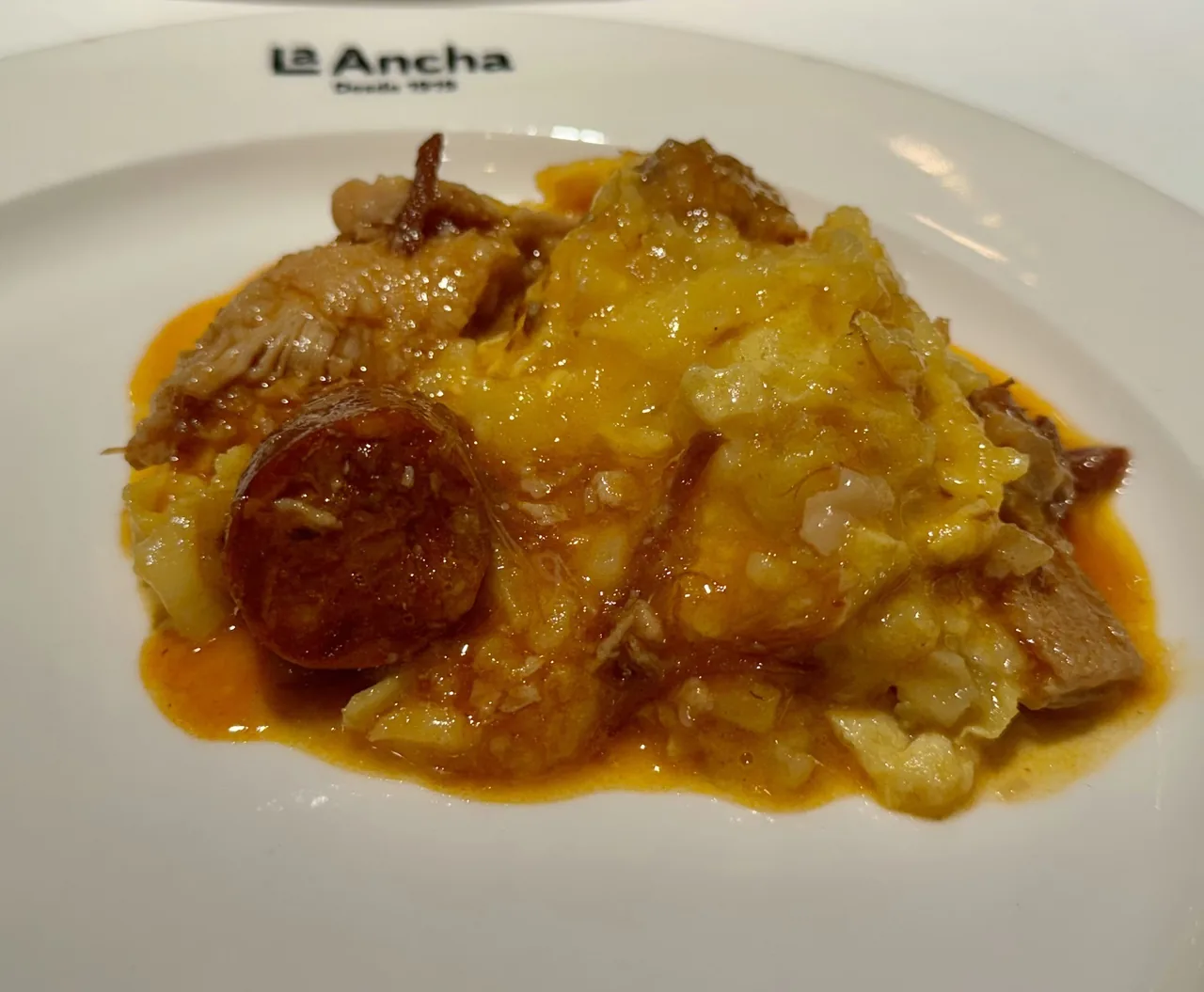 A plate of stewed tripe with a slice of chorizo in orange sauce on a white plate marked La Ancha