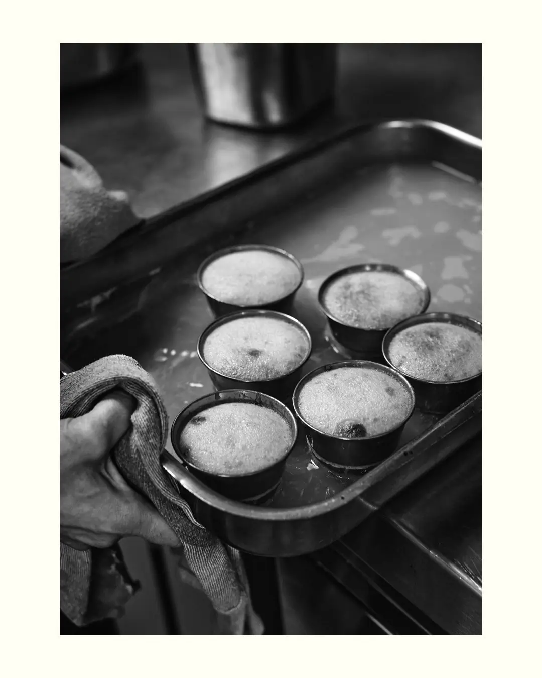 Black and white photo of a hand holding a tray of small metal cups filled with a foamy custard, likely flan.