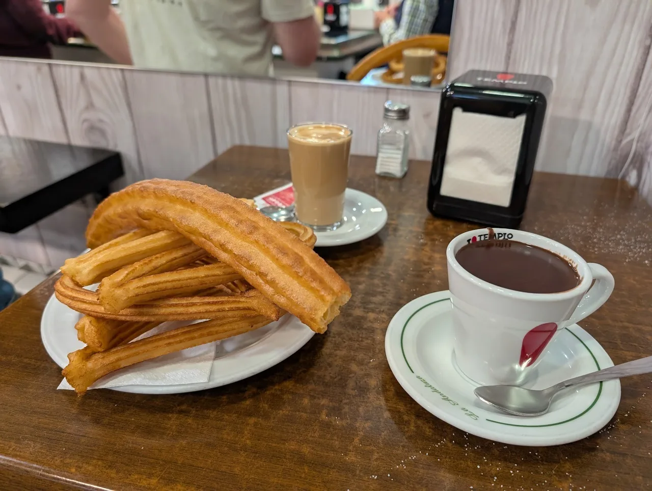 A plate of churros on a wooden table beside a cup of chocolate and a coffee