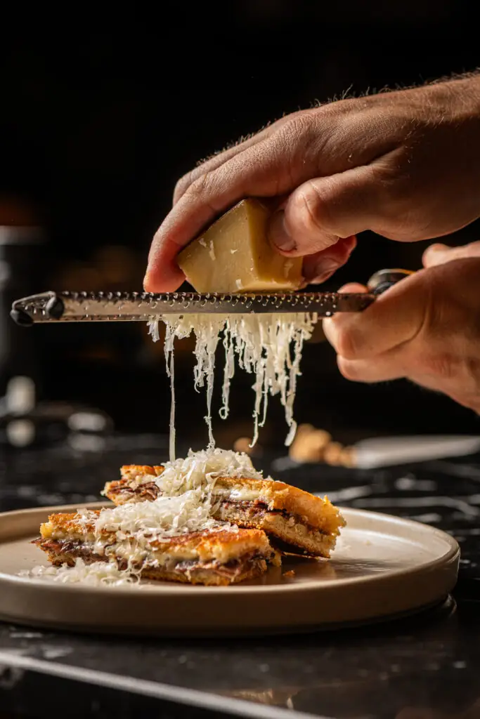 Hands grating a wedge of hard cheese over a toasted, filled sandwich cut into pieces on a pale plate against a dark background