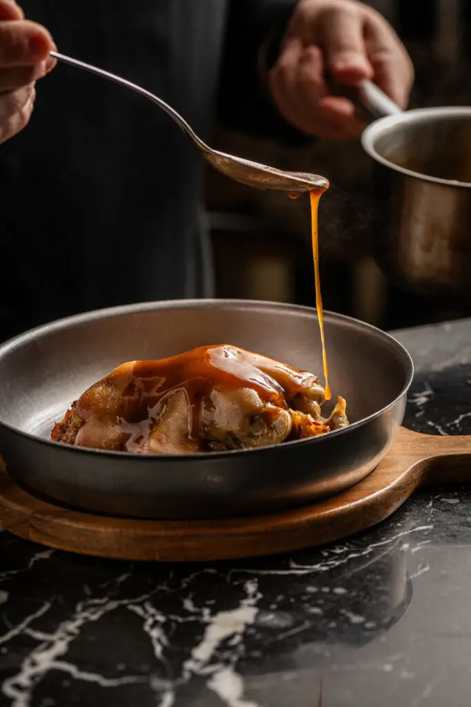 A braised pig's trotter or meat in a metal pan on a wooden board being spooned with glossy amber sauce on a black marble counter