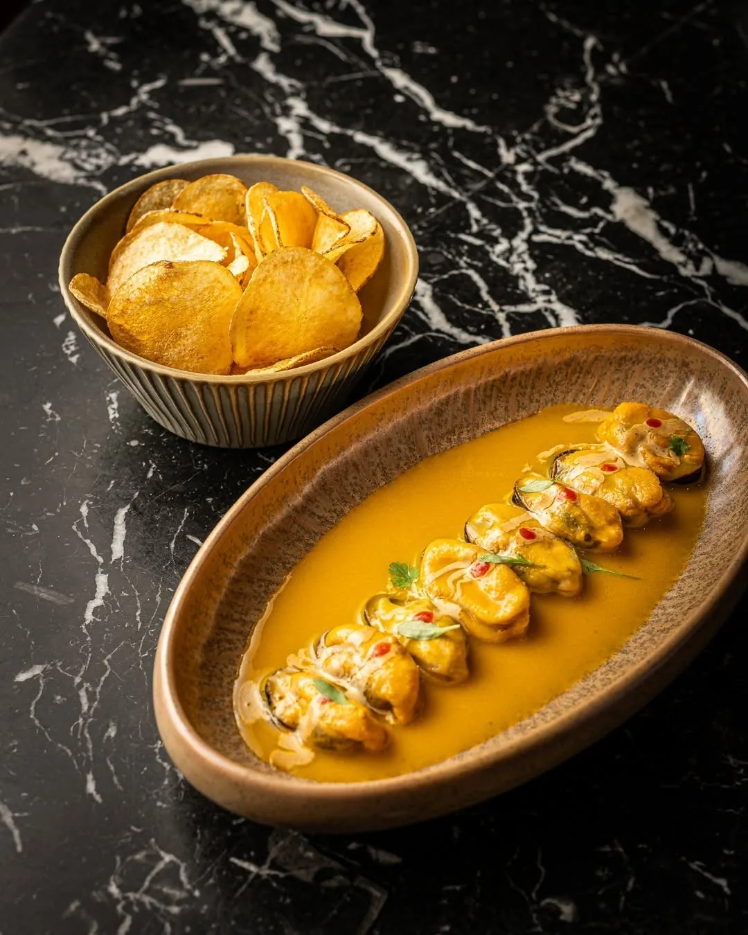 An oval bowl of a yellow sauced dish beside potato chips on black marble
