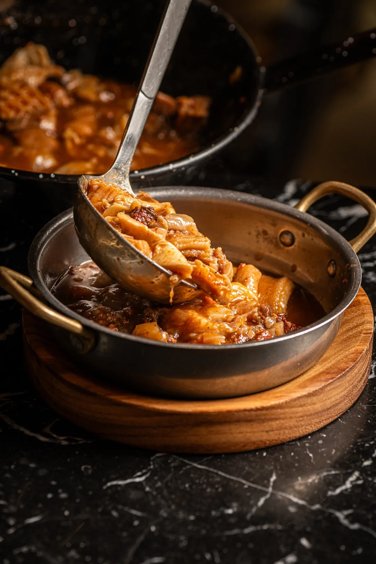 A ladle pouring stewed meat and sauce into a small metal pan on a wooden board