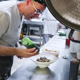 Chef plating a dark sauce over a dish in a white bowl in the kitchen