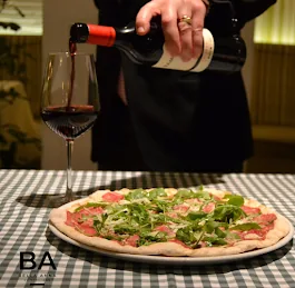 A pizza topped with arugula and prosciutto, wine being poured beside it