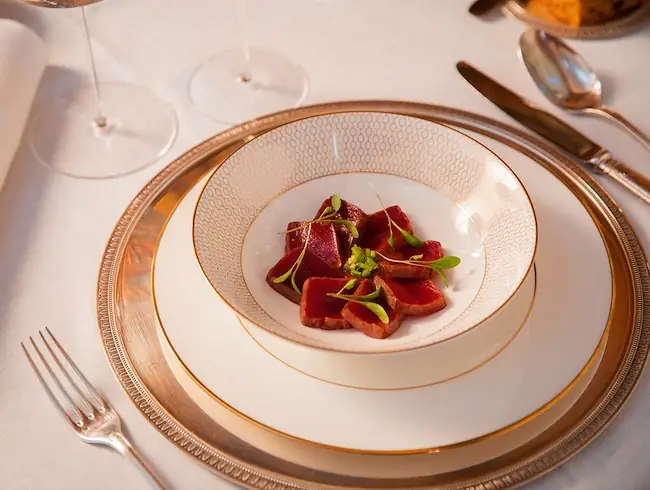 Slices of seared rare tuna with wasabi and microgreens plated in a gold-rimmed bowl on a silver charger