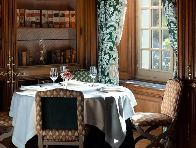 Elegant wood-panelled dining room with a set round table, wine glasses, a bookcase and patterned upholstered chairs by a window