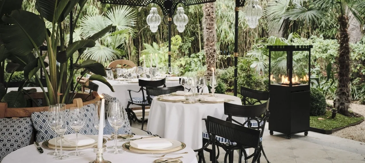 Tables set with white cloths, candles and glasses among palms in a garden terrace
