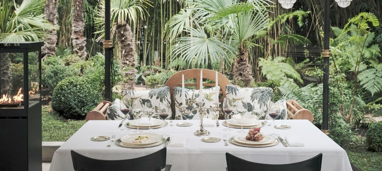 A garden dining table with plates and glasses set among palm trees and greenery