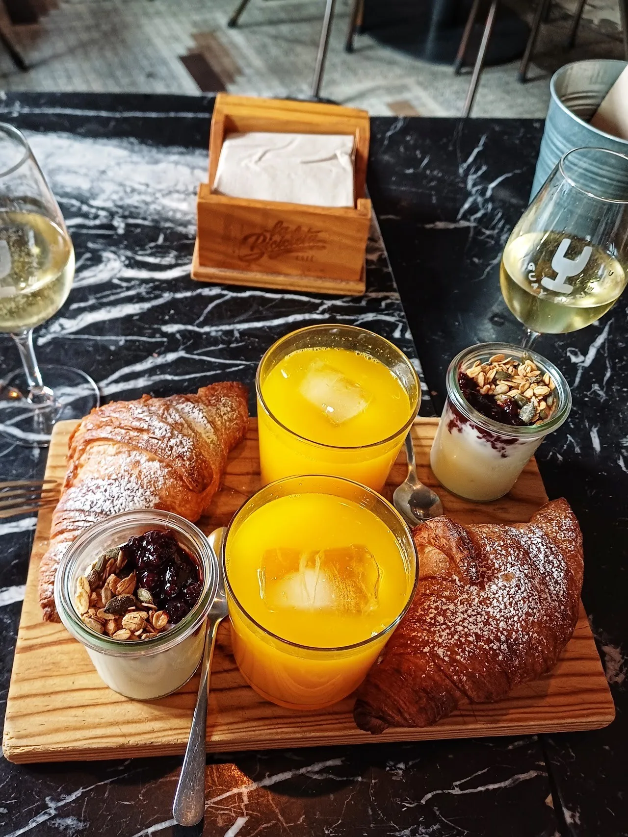 A wooden board with a croissant, two glasses of orange juice and two jars of yoghurt with granola