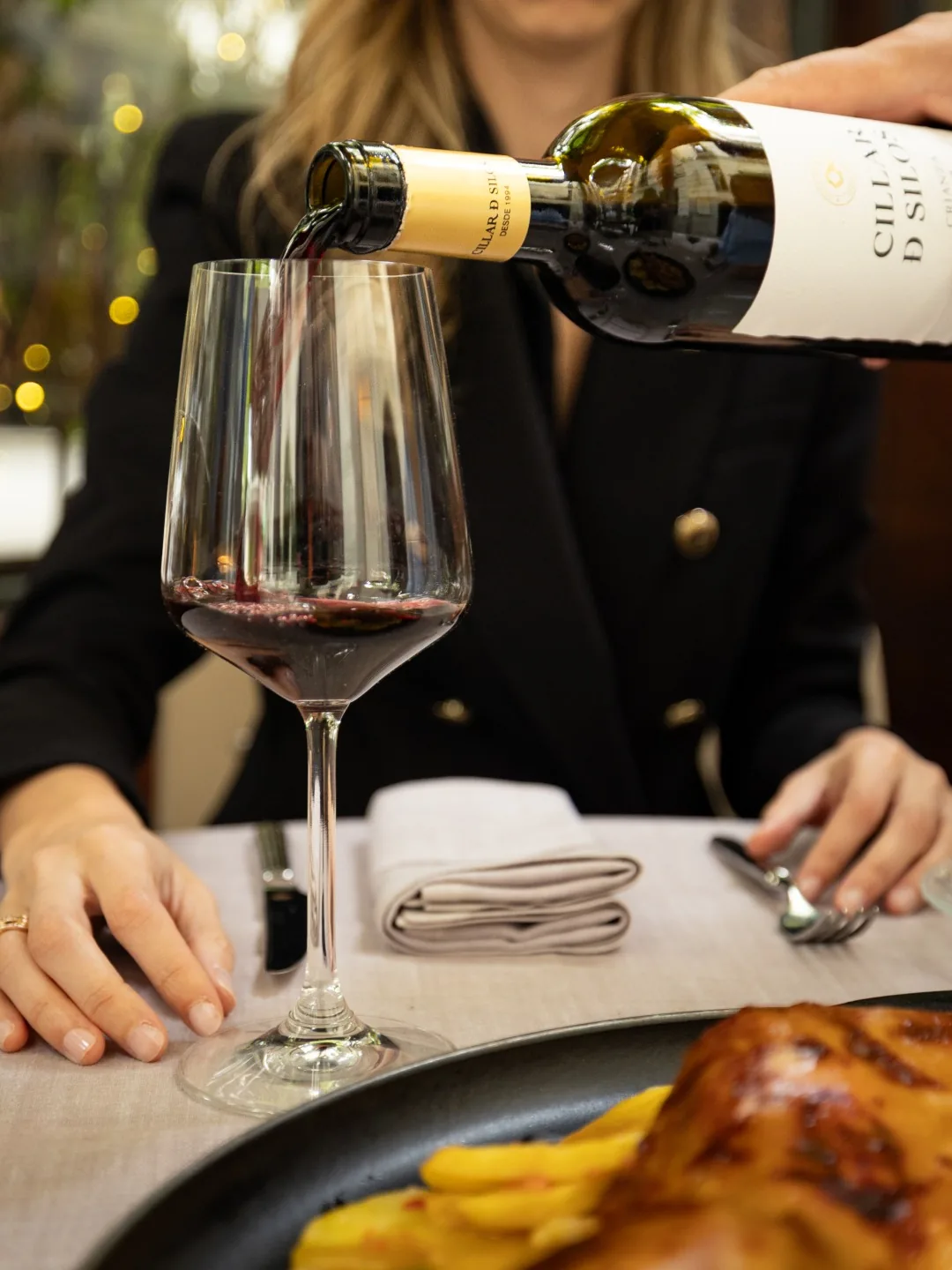 Red wine being poured into a glass at the table, with a roasted meat and potato dish visible in the foreground