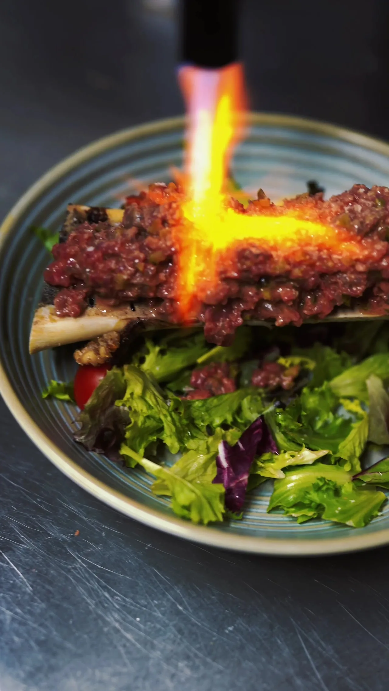 A dish being flambeed with a visible flame, salad and vegetables on the plate