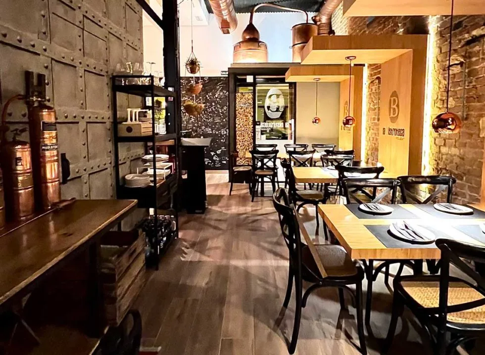 La Bistroteca's dining room with a riveted metal wall, copper vessels on a wooden ledge, bentwood chairs at laid tables, exposed brick and copper pendant lamps, the fingerprint-B logo mounted at the back