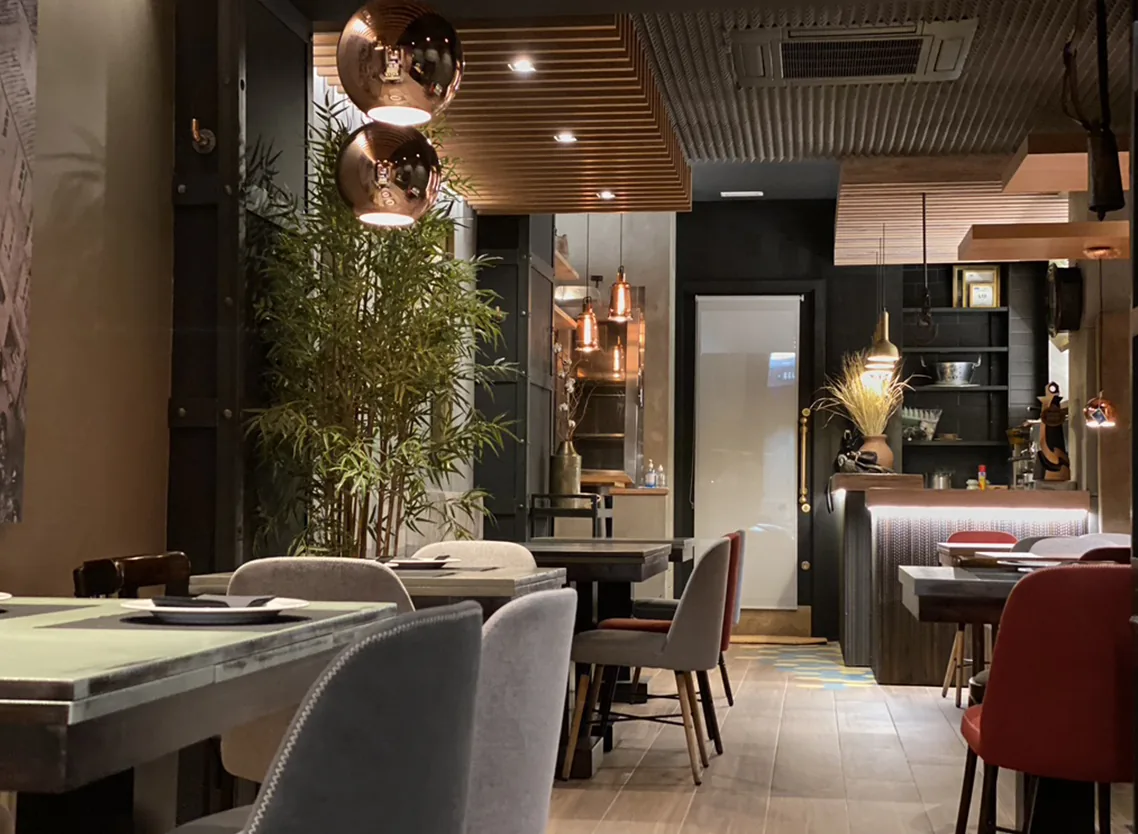 A second dining area with copper globe pendant lights, a tall bamboo plant, grey and burgundy upholstered chairs at laid tables, and a service counter at the back