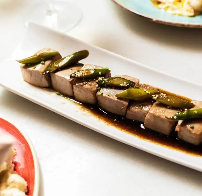 Seared tuna loin sliced and topped with grilled green padron peppers and sesame seeds in a soy glaze, plated on a rectangular white dish, at La Bodegueta Provença