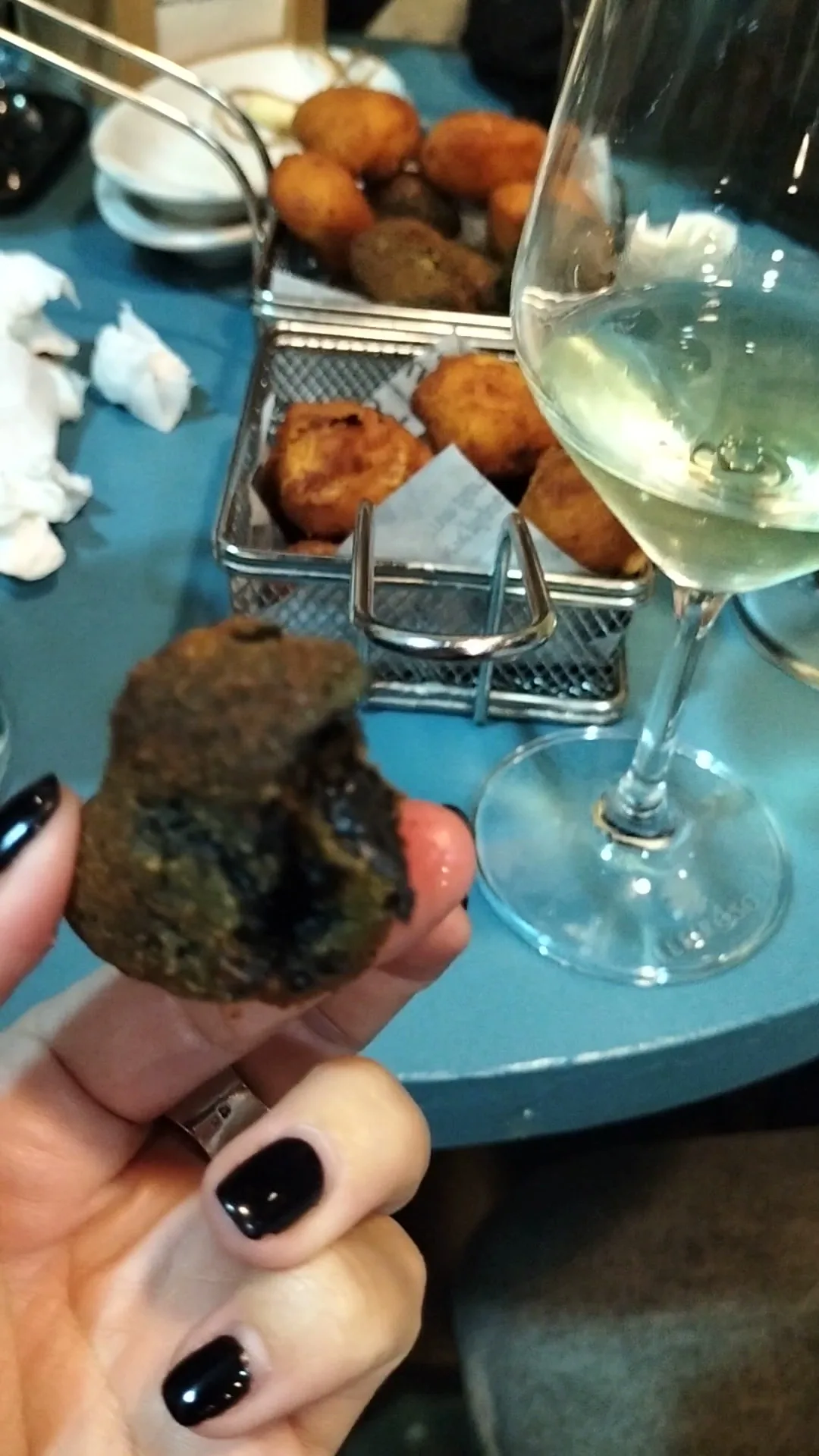 A hand holding a croqueta beside a plate of croquettes and a glass of wine