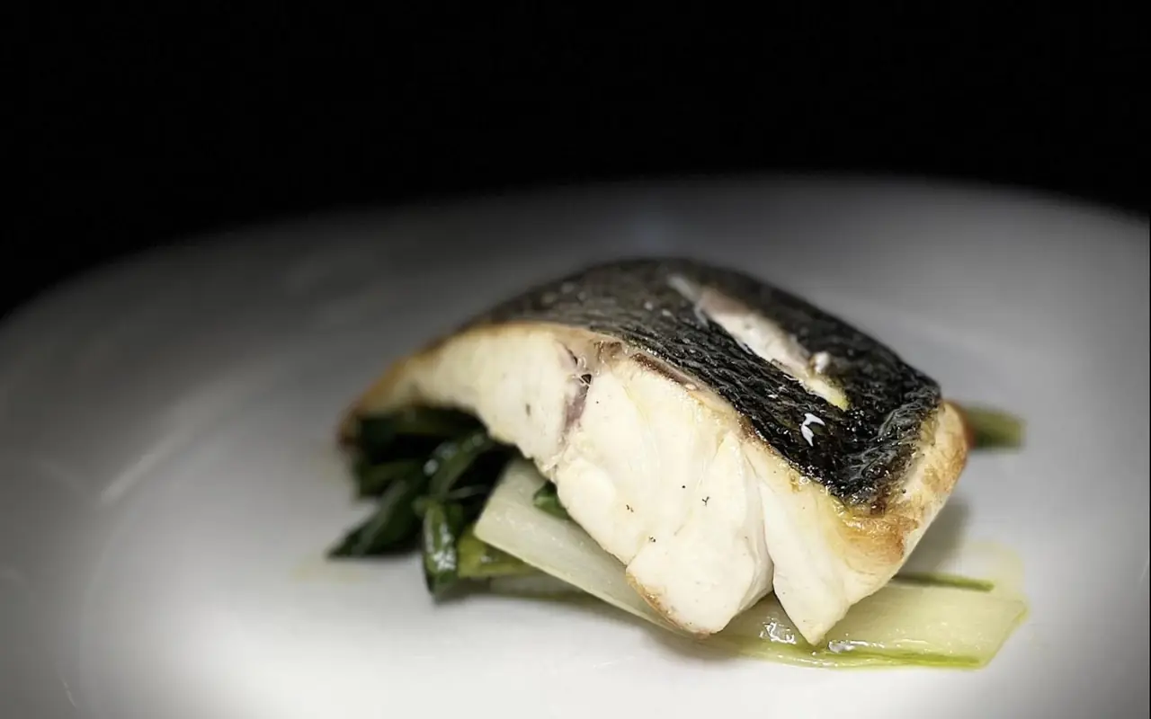 Crisp-skinned white fish fillet resting on sauteed greens with pale sliced vegetable on a white plate.