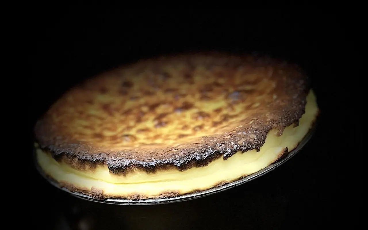 Whole baked cheesecake with a scorched brown top and pale custardy centre, tilted on a metal base against black.