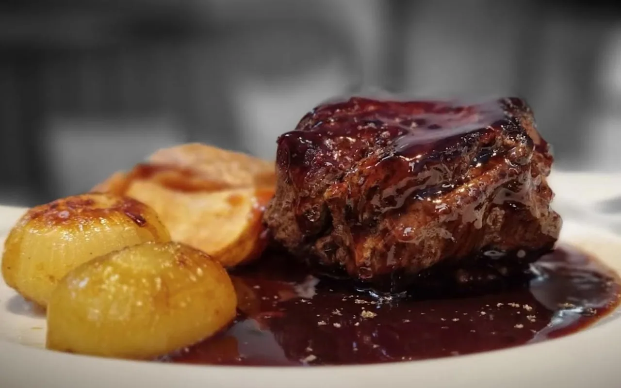 A piece of braised meat in dark sauce with two glazed onions on a white plate
