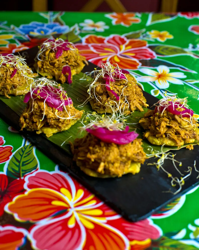 Six tostones topped with shredded beef, pickled red onion and sprouts, lined up on a slate board over a floral oilcloth