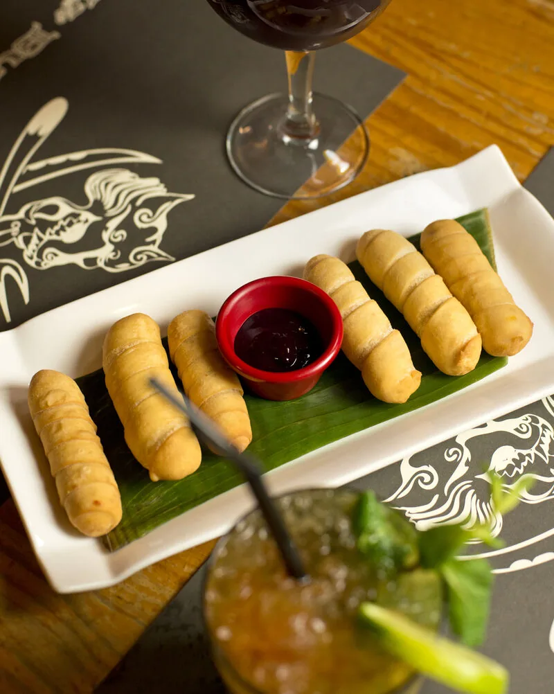 Row of tequenos with a dark dipping sauce on a white plate, a glass of red wine and an iced cocktail alongside, on a placemat printed with the venue's wolf line-art