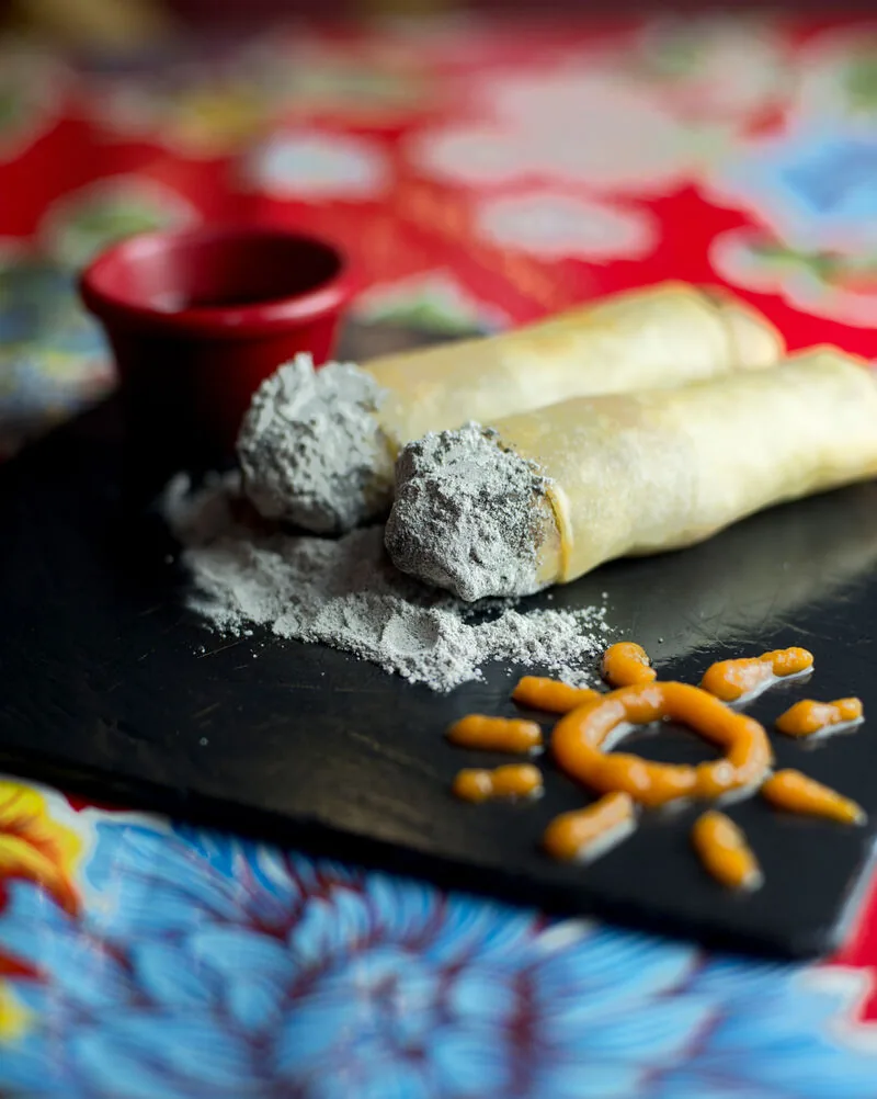 Two rolled crepes dusted with grey ash-coloured powder on a slate board, a sun drawn beside them in orange sauce