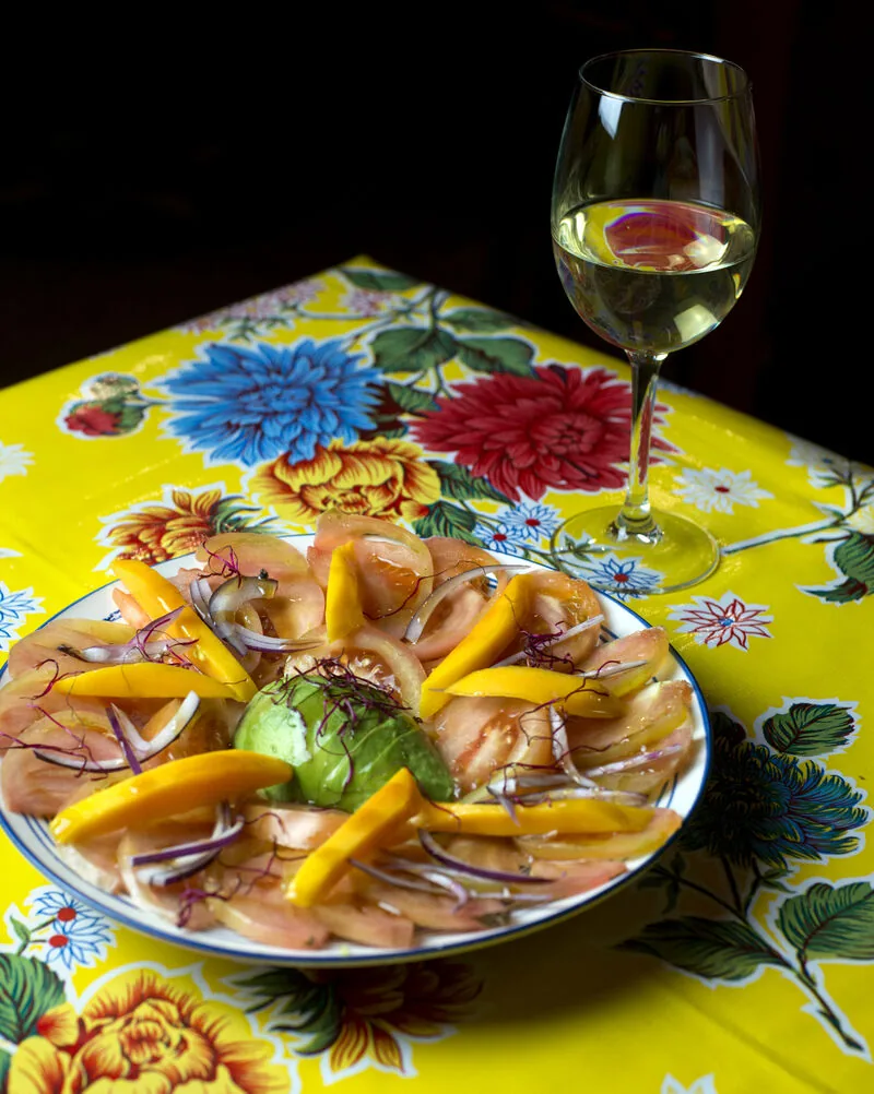 Sliced tomato and mango salad with red onion and an avocado quenelle on a rimmed plate, a glass of white wine behind, on a yellow floral oilcloth