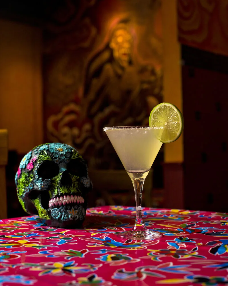 Salt-rimmed margarita with a lime wheel beside a painted sugar skull, on a pink floral tablecloth in the muralled room