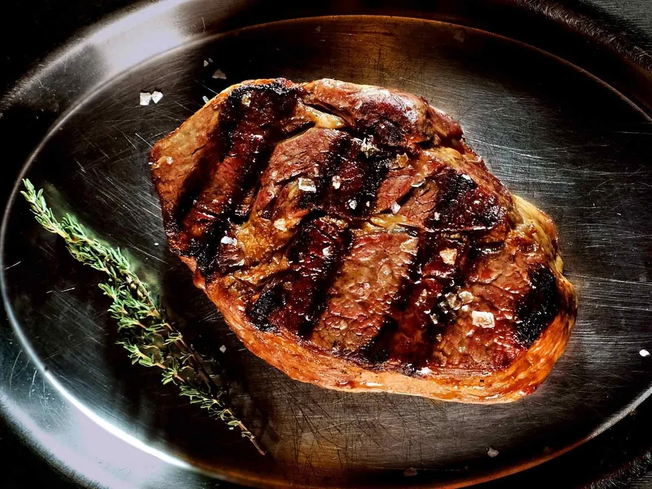 A grill-marked steak on a dark plate with a sprig of thyme