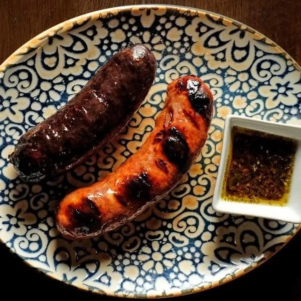 A grilled blood sausage and a chorizo on a patterned plate with a small dish of herb sauce