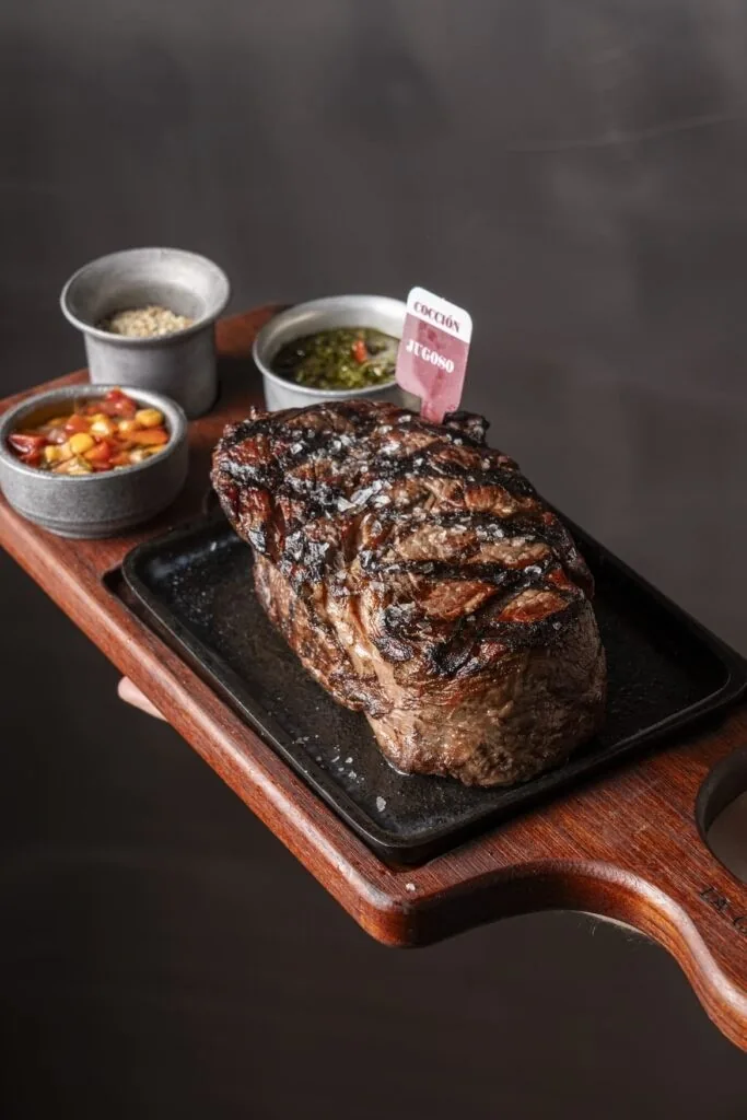 A thick grilled steak on a cast-iron plate on a wooden board with side sauces