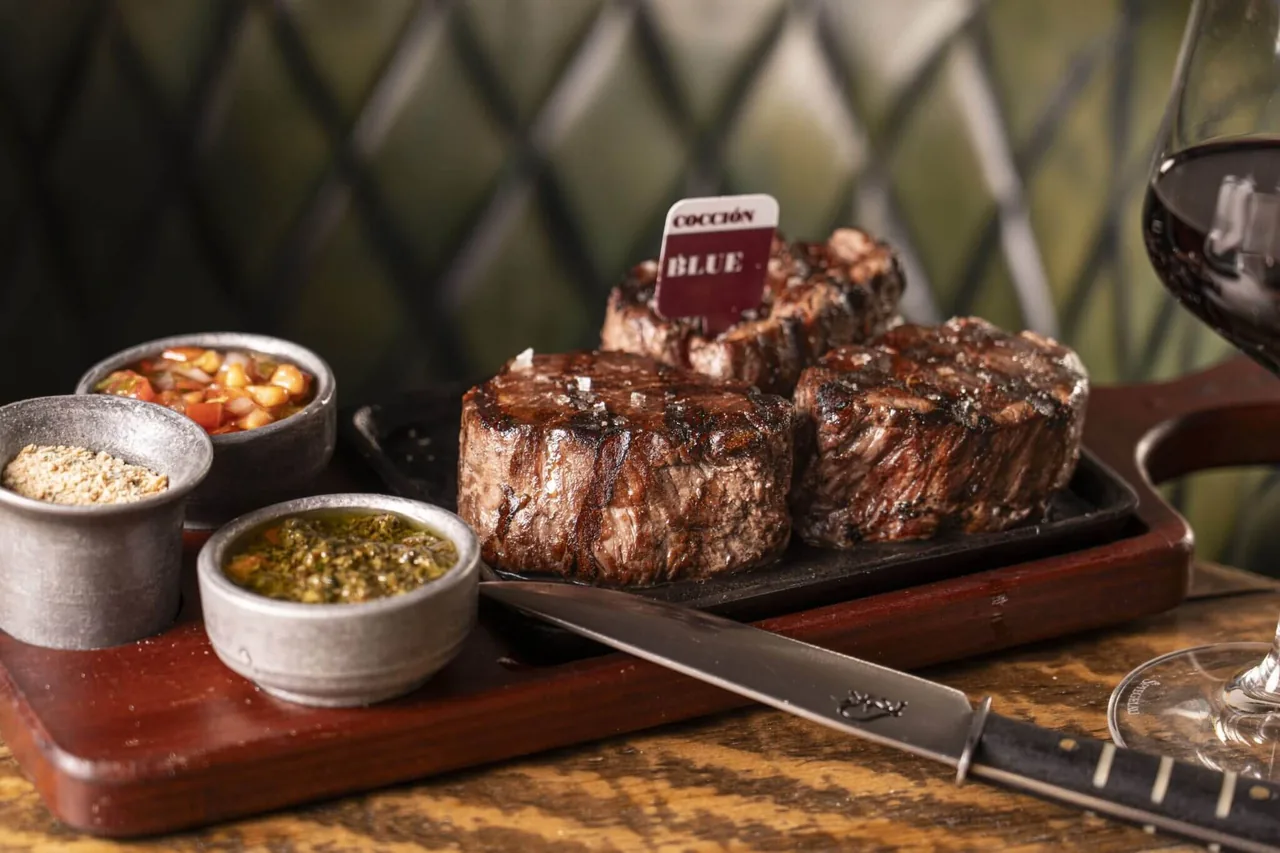 Three grilled beef medallions on a cast-iron plate flagged 'COCCION BLUE', with chimichurri, salsa criolla and a dry rub in metal pots on a wooden board, a glass of red wine at the edge