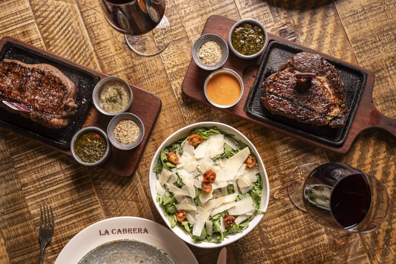 Overhead spread of two steaks on cast-iron plates with chimichurri and sauce pots, a rocket and parmesan salad and two glasses of red wine, a plate marked LA CABRERA in the corner